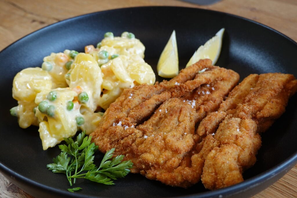 Gebackener Karpfen mit goldbrauner Panier und cremigem Erdäpfel-Mayonnaise-Salat auf schwarzem Teller