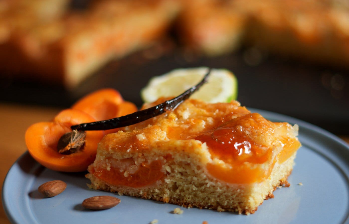 Ein Stück Marillenkuchen mit glänzenden Marillen, Goldhaube und Vanilleschote auf einem Teller.