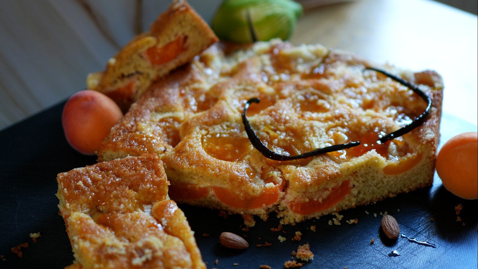 Marillenkuchen mit Anschnitt, Goldhaube und sichtbaren Marillenstücken auf einer dunklen Platte.