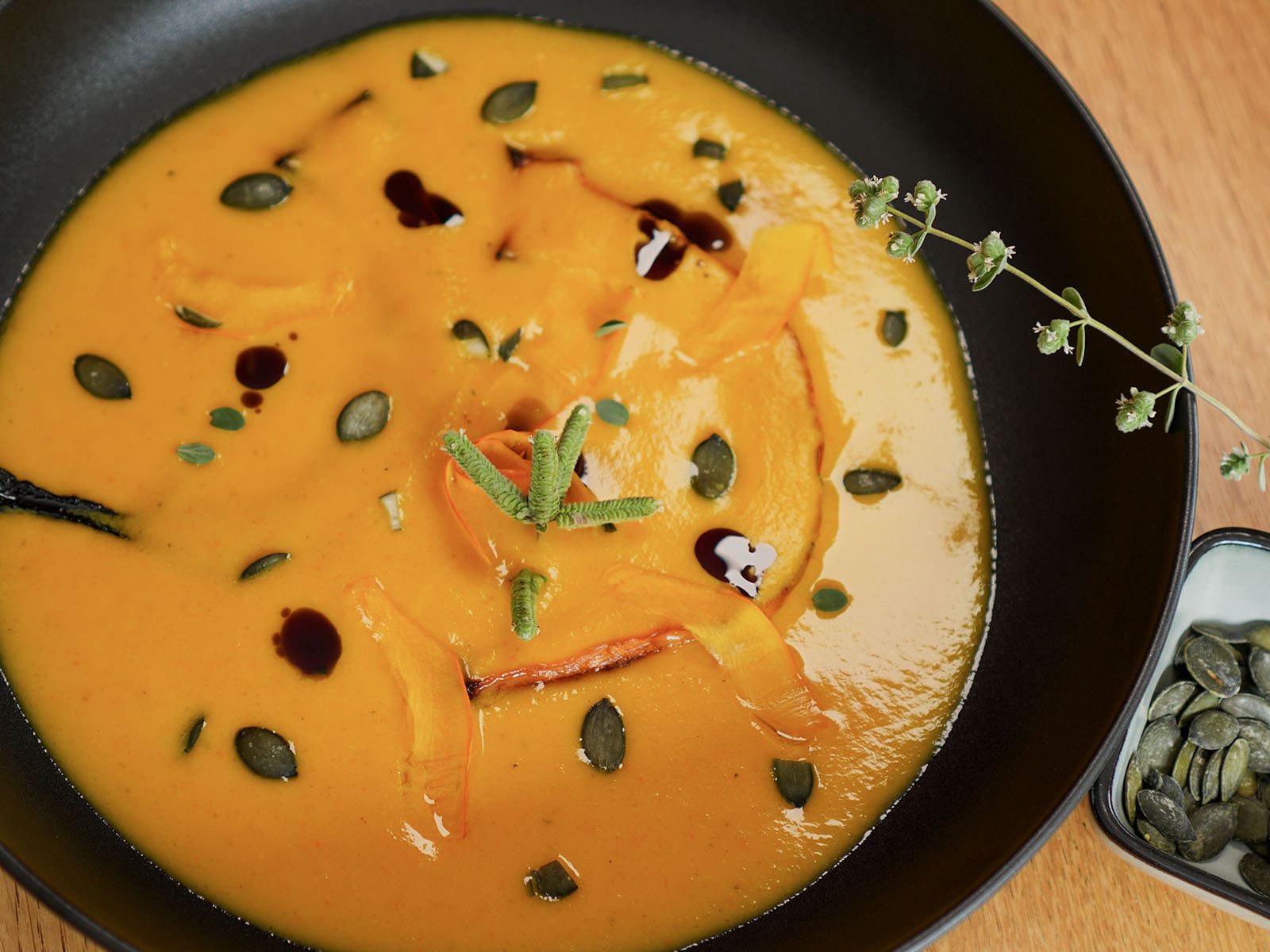 Kürbiscremesuppe von oben in schwarzer Schale, mit Ofenkürbis-Streifen, Kernen und Majoran, daneben eine Schale Kürbiskerne.