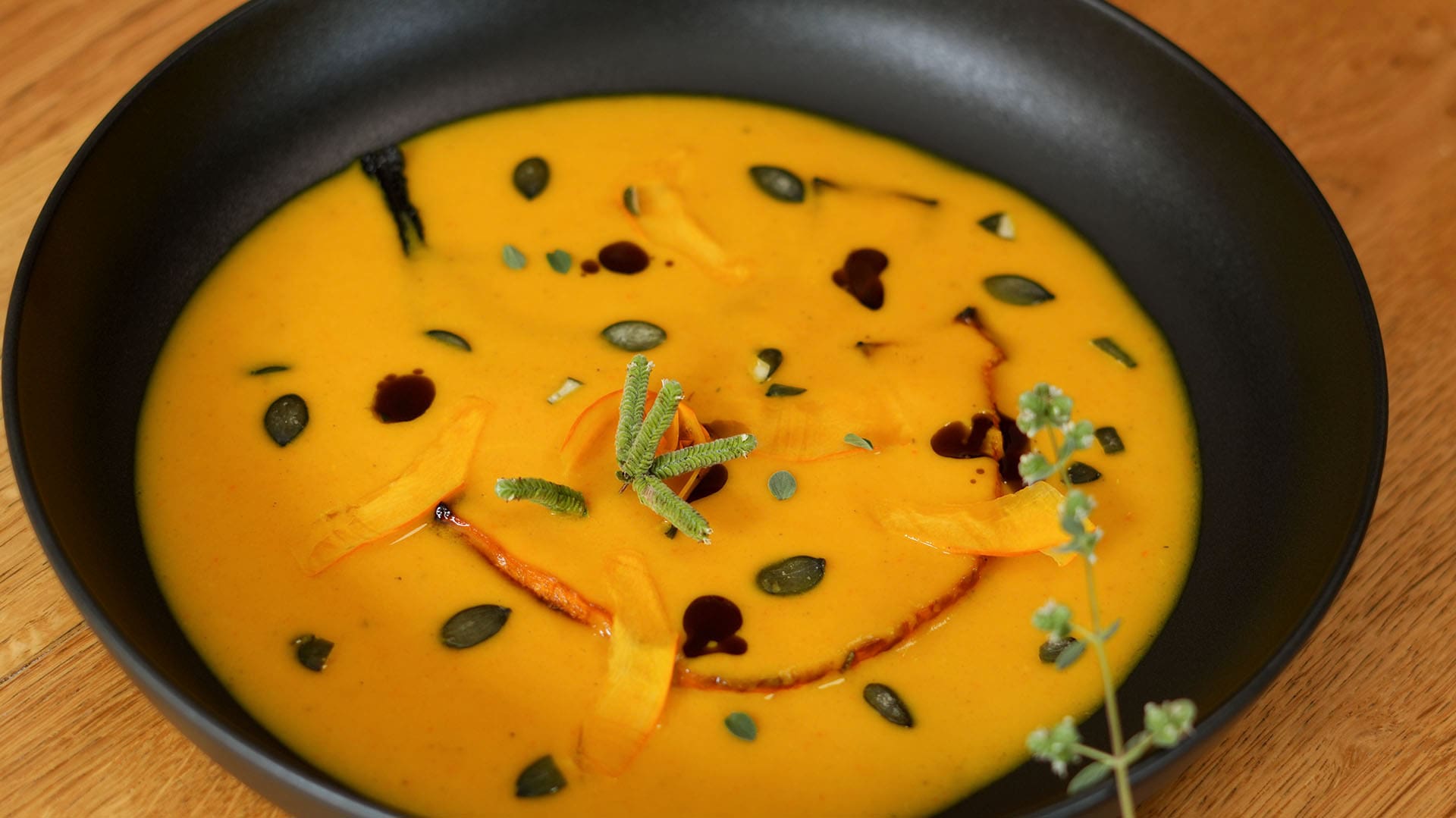 Kürbiscremesuppe in schwarzer Schale, mit Kernöl-Tropfen, gerösteten Kürbiskernen, rohen Kürbisstreifen und Majoran.