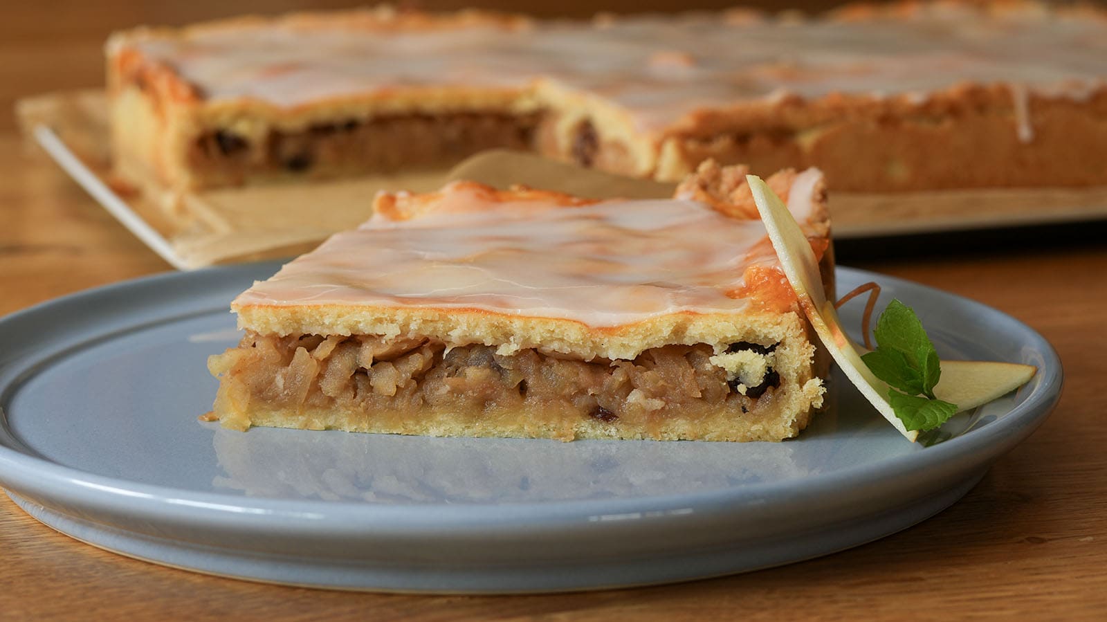 Gedeckter Apfelkuchen mit Mürbteig und Zuckerguss, ein Stück angeschnitten auf blauem Teller, dahinter der ganze Kuchen
