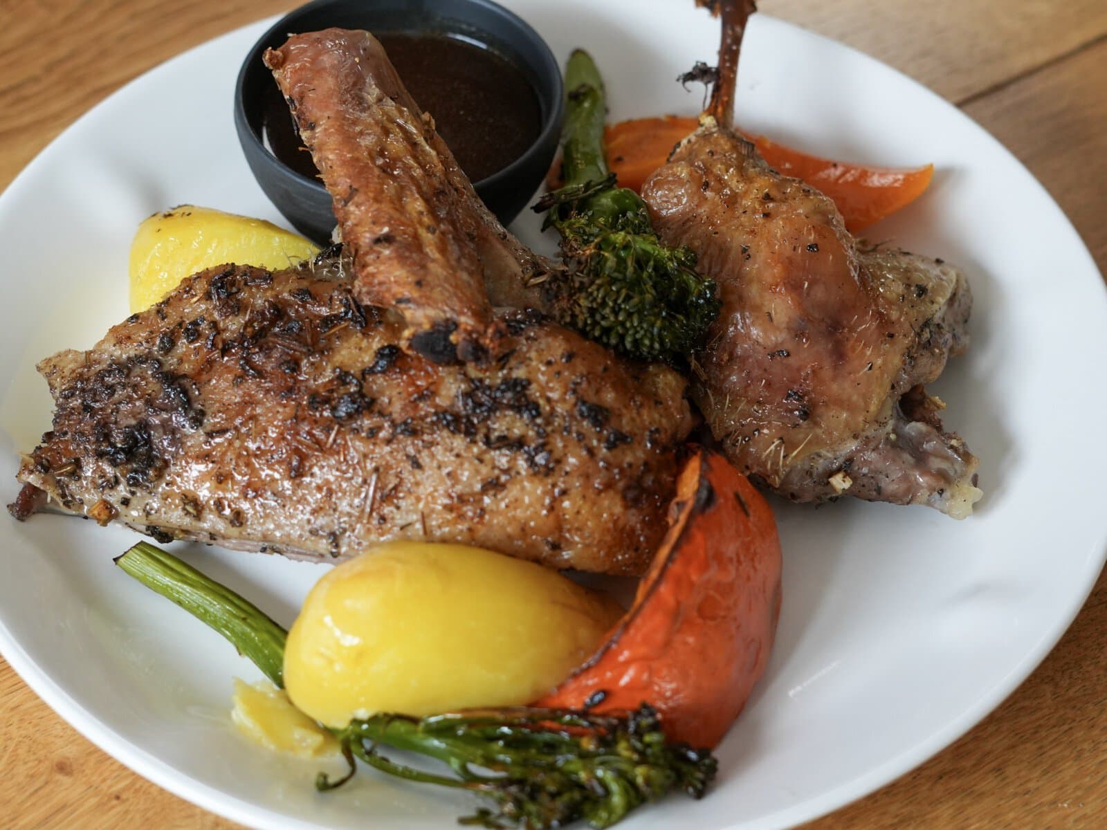 Portion knusprige Ente mit Keule und Brust auf weißem Teller, dazu Kartoffeln, Kürbisspalte, Stangenbrokkoli und ein Schälchen Entensoße