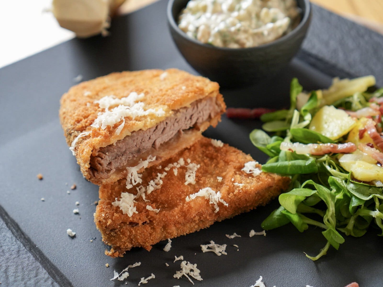 Alt-Wiener Backfleisch nah: goldbraune Panier, angeschnitten, mit frisch geriebenem Kren, dahinter Vogerlsalat mit Speck